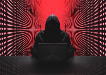 Anonymous hacker in front of his computer with red light wall backgroundAnonymous hacker in a black hoody with laptop in front of a code background with binary streams cyber security concept